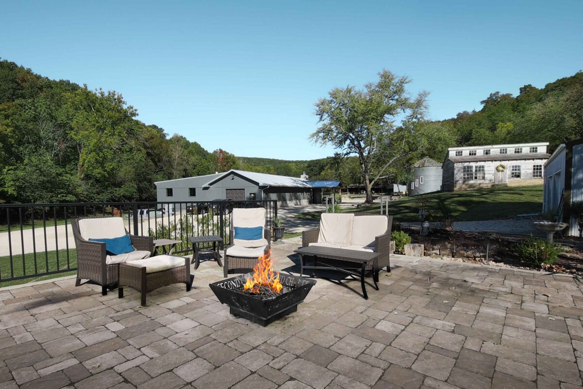Patio furniture outdoors on a stone tiled surface with a lit fire pit in the middle.