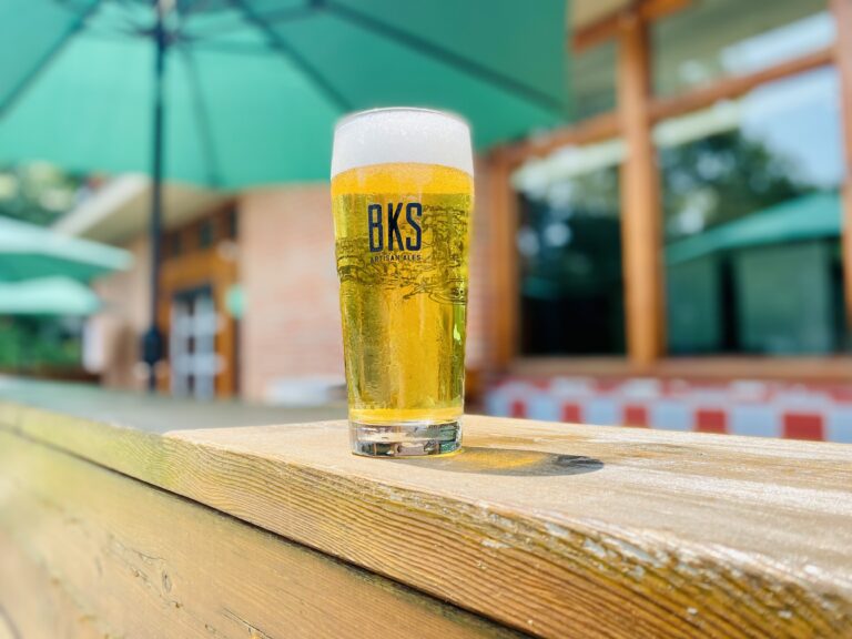 A tall glass of beer with a BKS logo sitting on a wooden table.