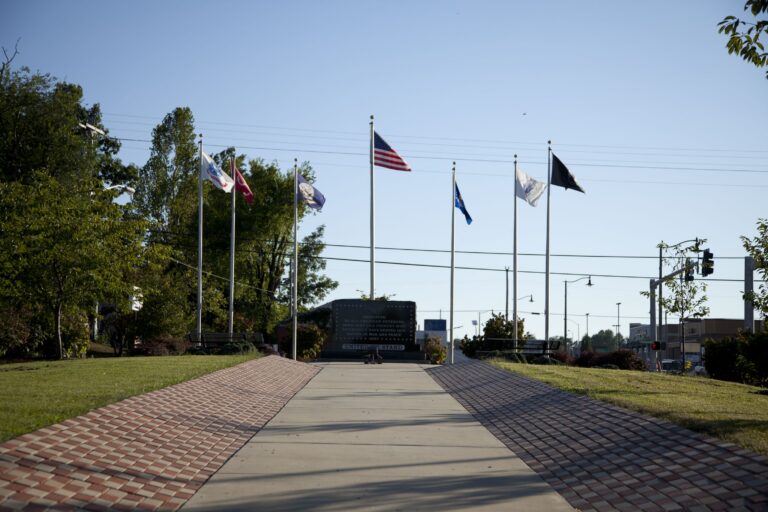 A concrete path lined with patterned brickwork leads to a black granite veterans memorial monument. The site features a central American flag surrounded by six other service and state flags, set in a park with green grass and mature trees under a clear sky.