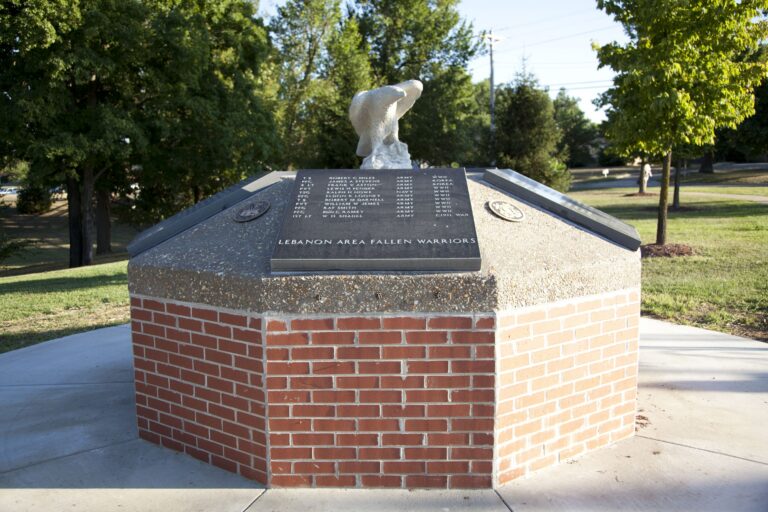 A multi-sided, red brick and concrete memorial topped with a white eagle statue. Dark granite plaques inscribed with "LEBANON AREA FALLEN WARRIORS" list the names and military branches of local service members. It is located outdoors, surrounded by grass and trees.