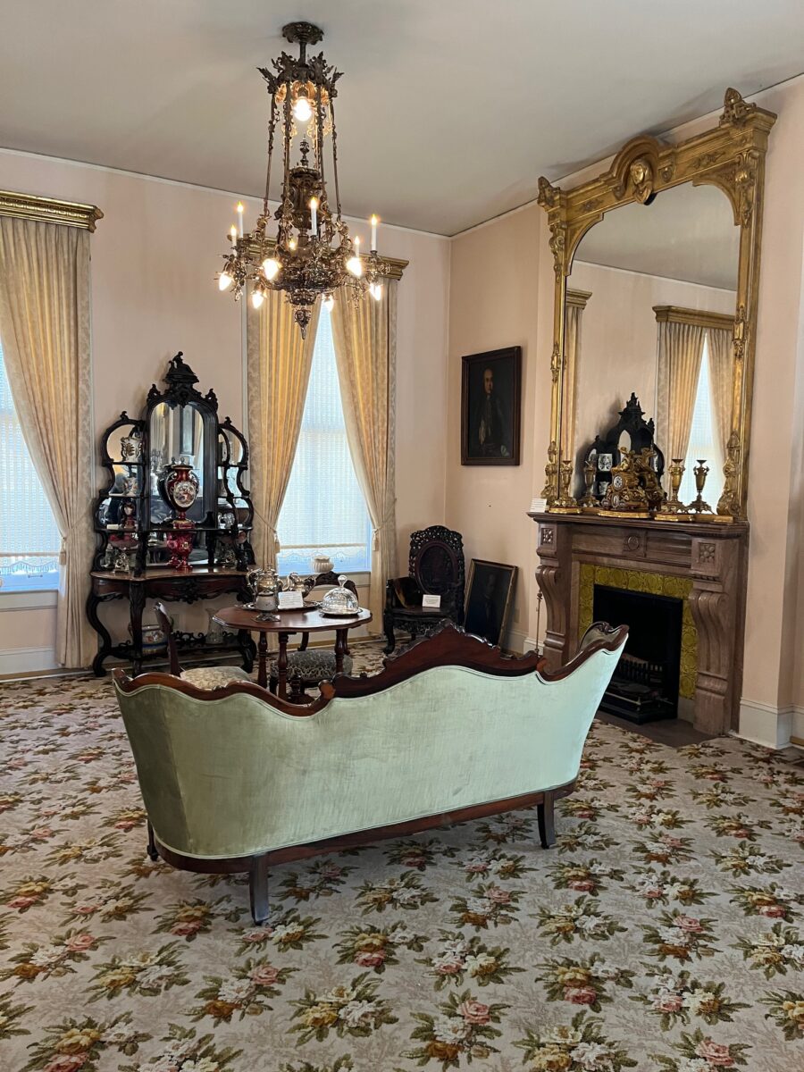A Victorian-era parlor with a mirror, chandelier and love seat.