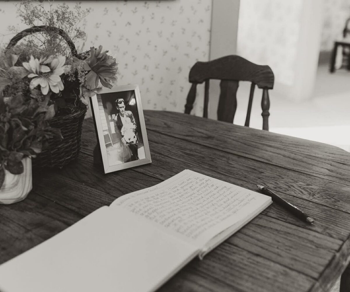 A black and white photo of a guest book and pen on a table in front of a framed photo and flowers.