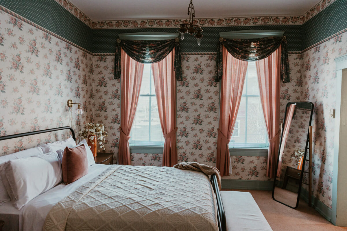 A wallpapered room with a made bed, a rug and two windows with curtains letting the sunlight in.