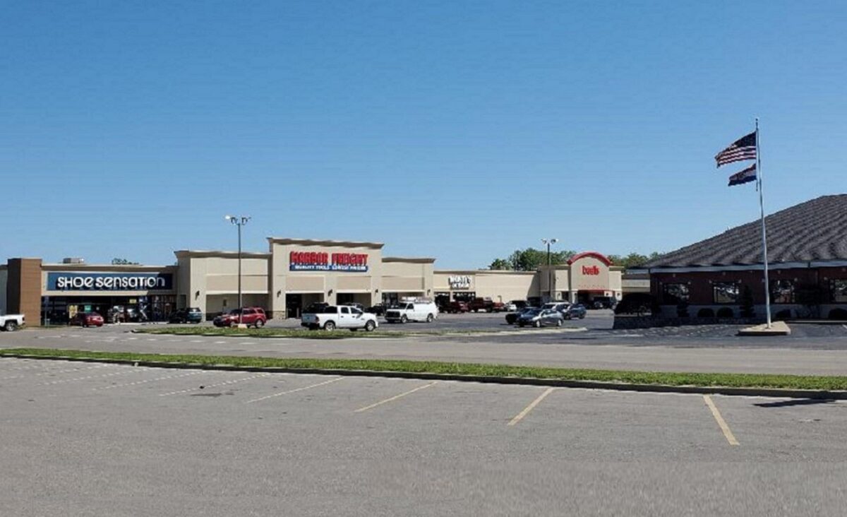 A view of a shopping center from the parking lot, showing storefronts, cars and raised flags.
