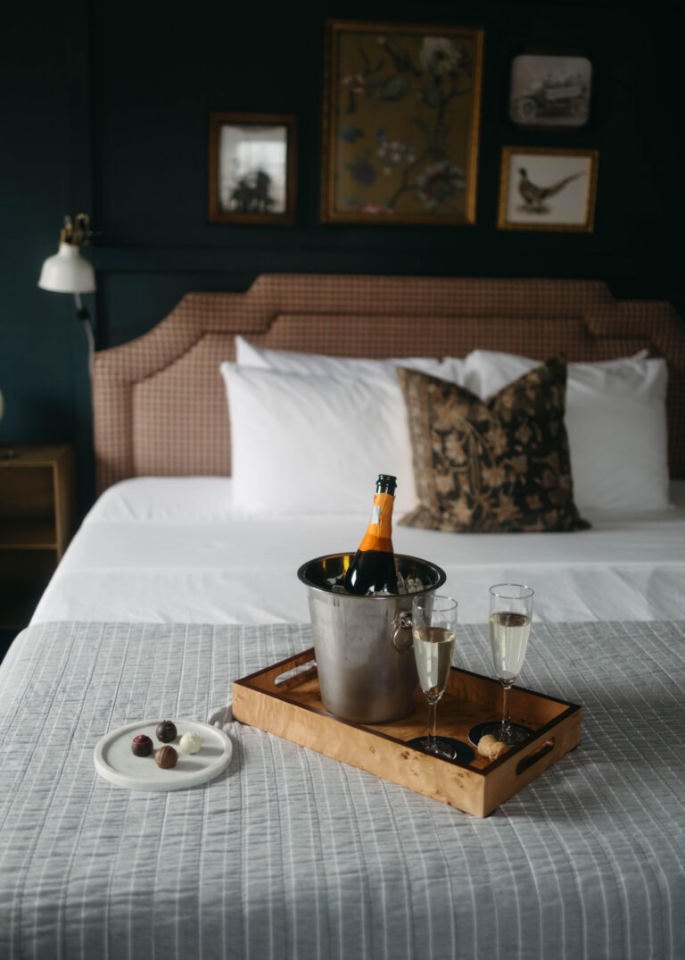 A champagne bucket, flutes and chocolate truffles sit on a tray on a cozy bed with white linens and a berry red gingham headboard.