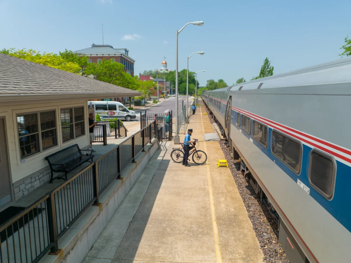 An aerial shot of a train station, with a grey roof and lighter siding, and its platform and a train with a person on a bike in between.