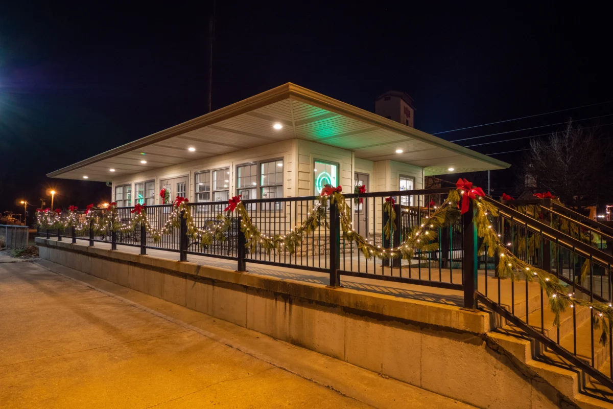 A night time shot of a train station, with a grey roof and lighter siding, and its platform railing adorned in Christmas hangings.