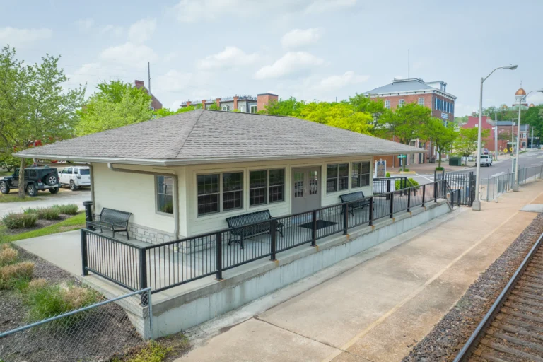 An aerial shot of a train station, with a grey roof and lighter siding, and its platform and tracks in front.