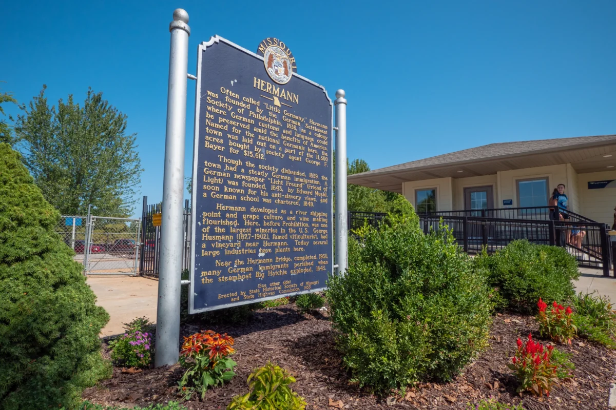 A large metal placard outside of a train station amid bushes and other plant life.