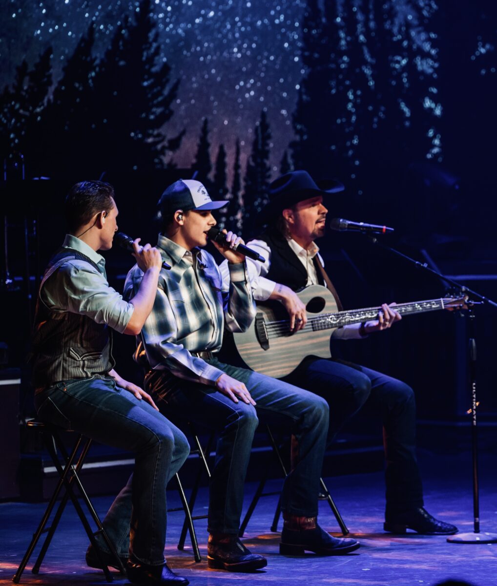 Three performers on stage before a colorful background.