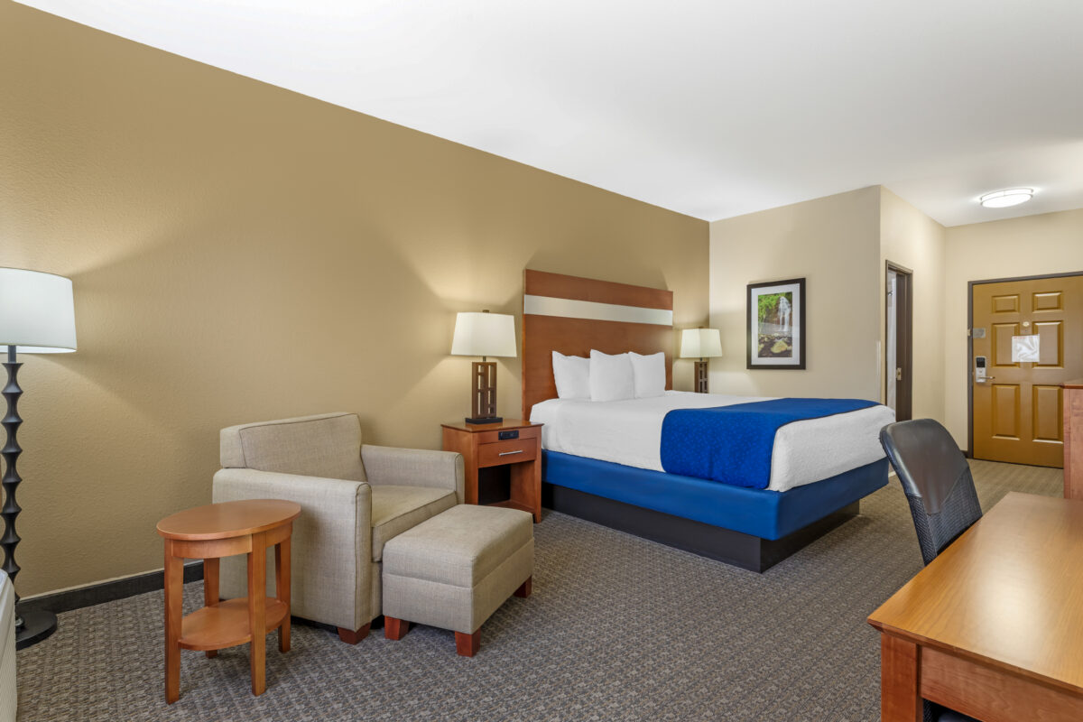 A furnished hotel bedroom with a bed, a sitting area, a desk and a television and blue tones.