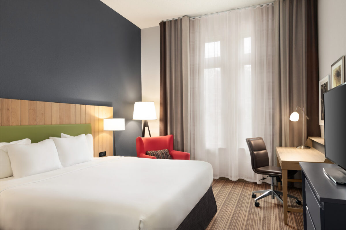 The interior of a hotel room with a bed, desk and tall windows.