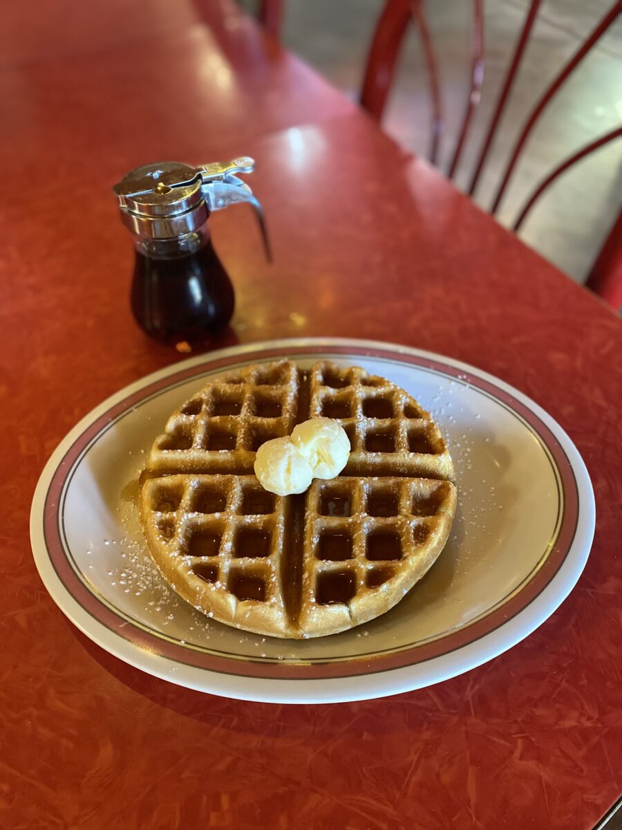 A waffle on a plate with butter on top, syrup on the side and a red table underneath.