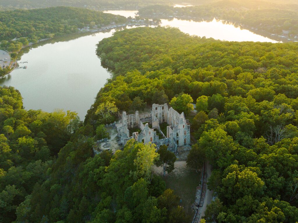 Castle ruins sit atop a bluff surrounded by lush green trees and flanked by a body of water. 
